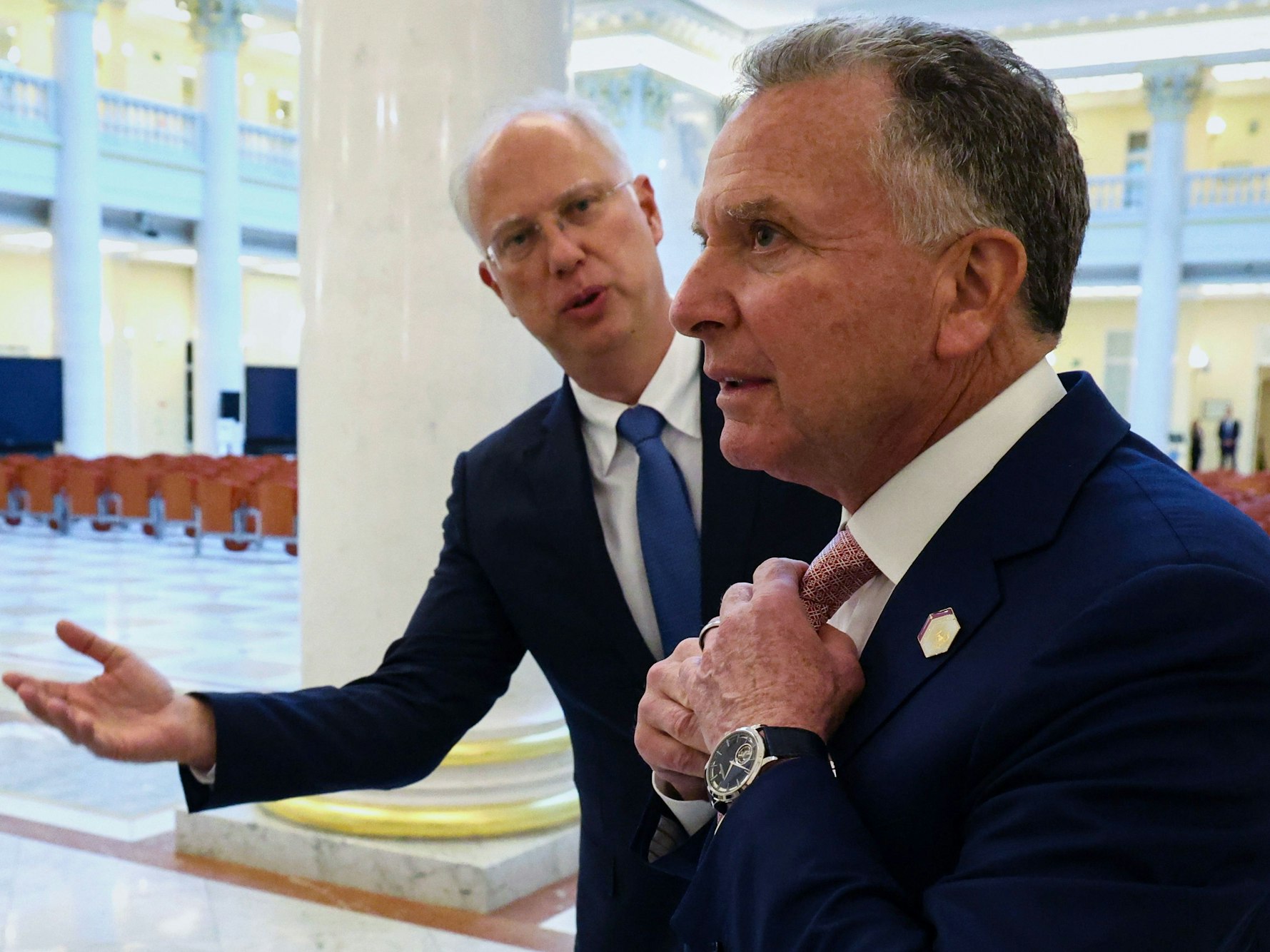 Ein Mann macht mit seinem rechten Arm eine ausladende Geste gegenüber einem anderen Mann, der sich die Krawatte zurechtrückt.