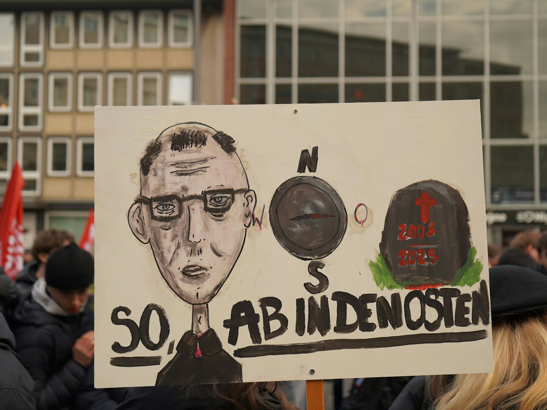 Eine Karikatur von Friedrich Merz, die Aufschrift „Ab in den Osten“ und ein Grabstein sind auf dem Schild zu sehen.