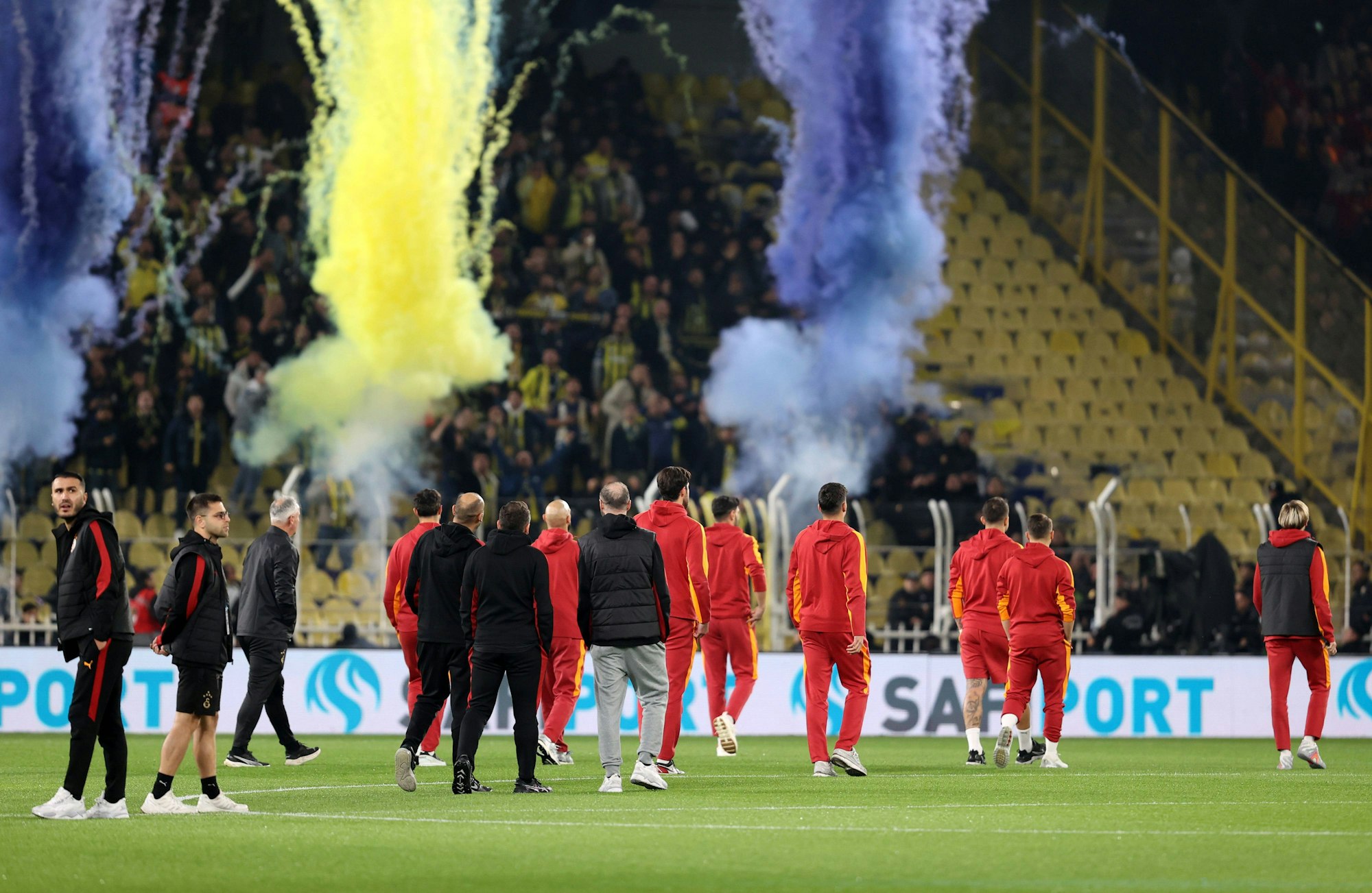 Galatasaray-Spieler stehen auf dem Rasen, im Hintergrund zünden Fenerbahce-Fans Rauchbomben.