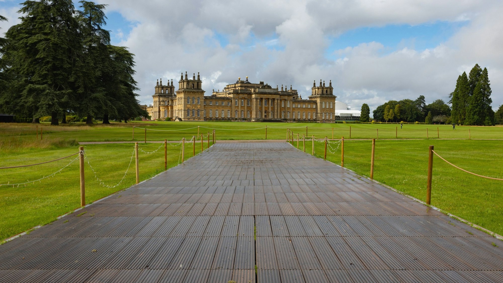 Ein Holzsteg führt zu Blenheim Palace, umgeben von Grünflächen, Bäumen und bewölktem Himmel
