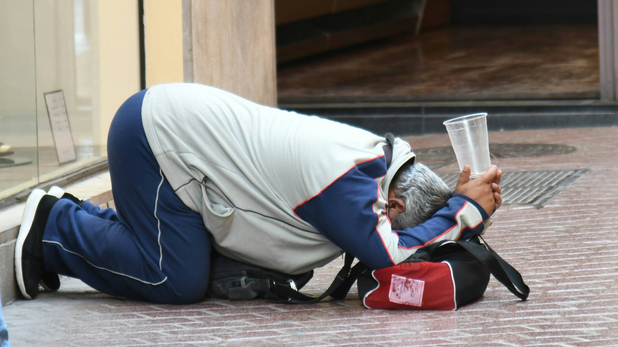 Ein alter Mann bettelt mit einem Becher kniend auf dem Boden in Palma de Mallorca