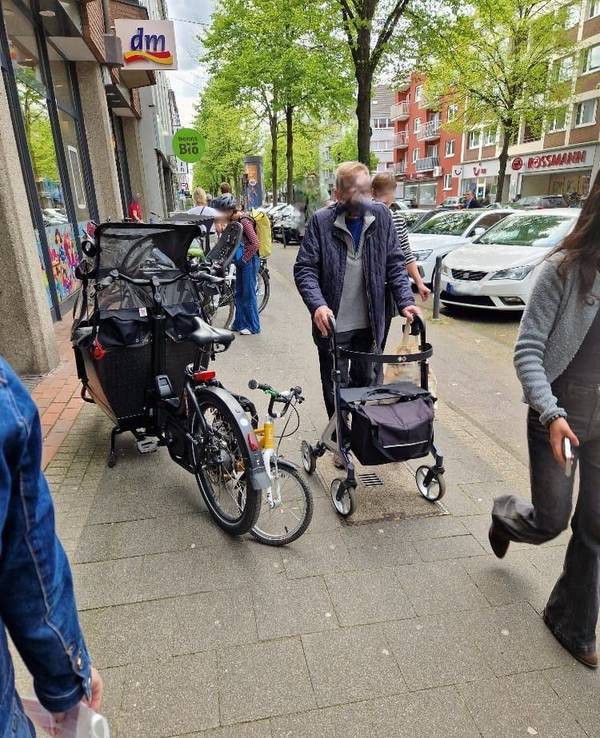 Ein Mann mit Rollator ist auf dem Gehweg unterwegs und muss an einem geparkten Lastenrad, das den halben Weg blockiert, vorbei.