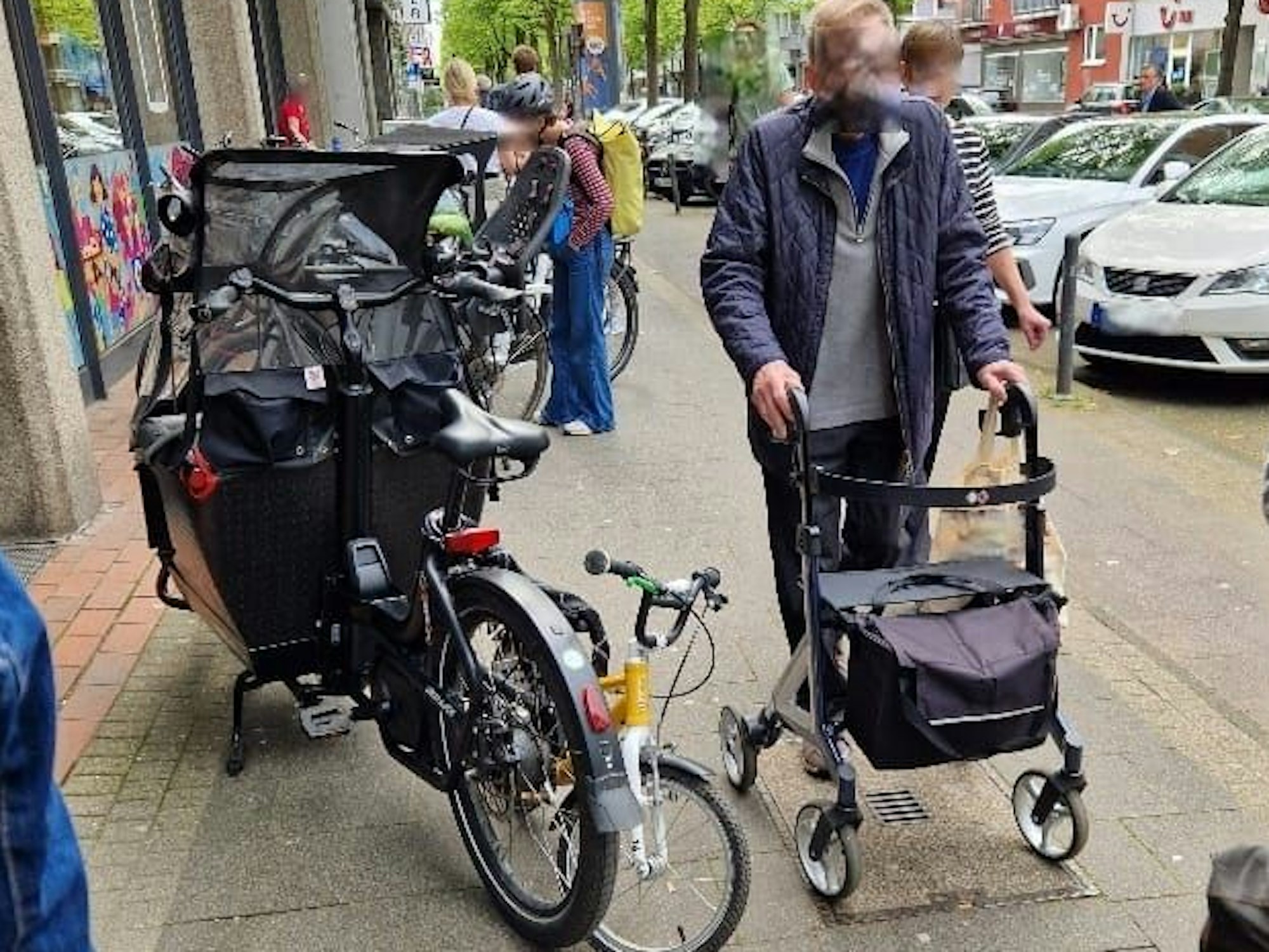 Ein Mann mit Rollator ist auf dem Gehweg unterwegs und muss an einem geparkten Lastenrad, das den halben Weg blockiert, vorbei.
