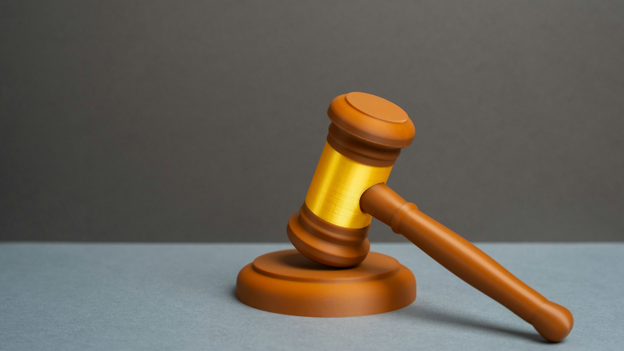 Ein brauner Richterhammer mit goldenem Band auf hellgrauer Fläche vor dunklem Hintergrund