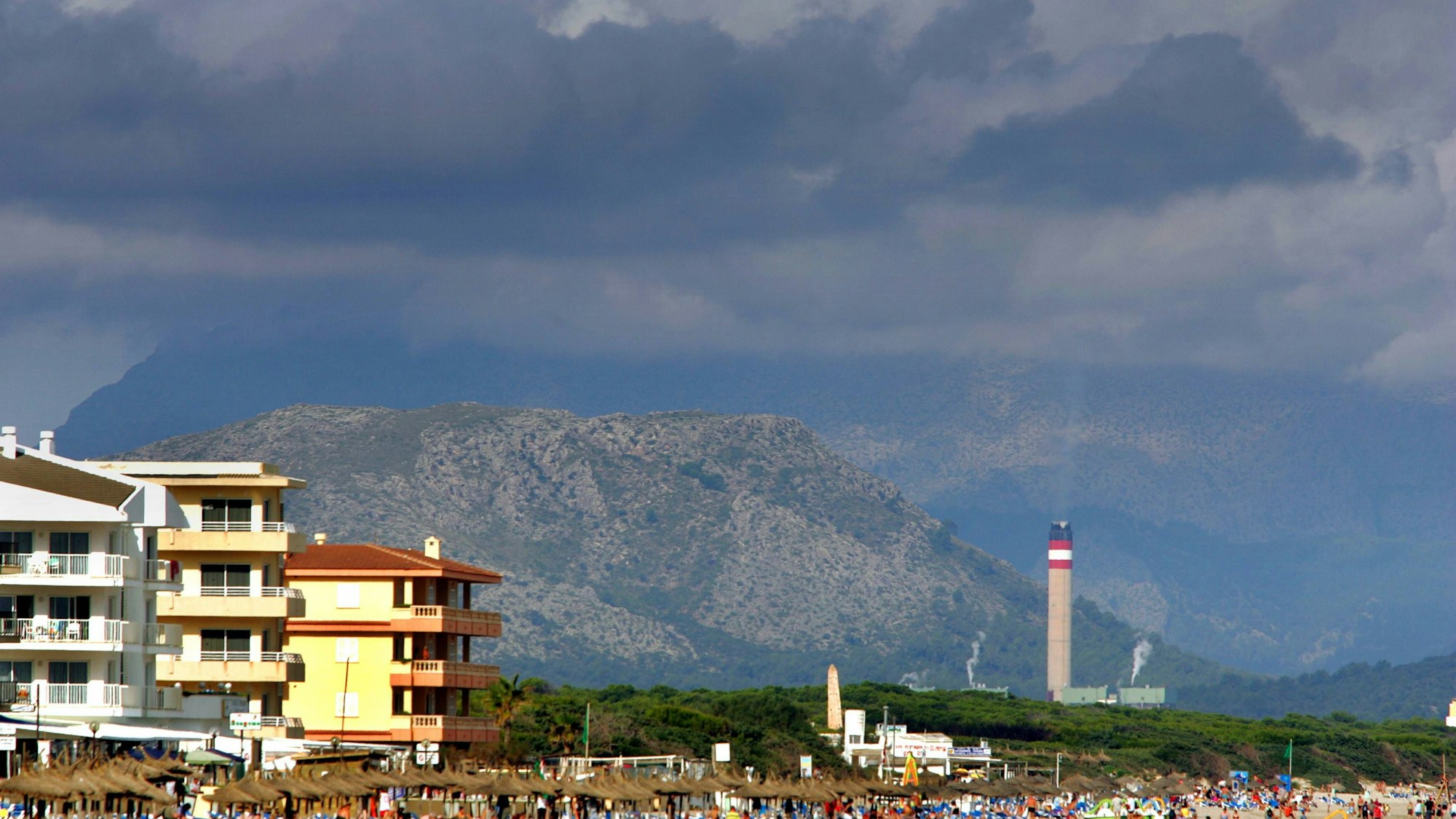 Belebter Strand von Can Picafort auf Mallorca unter dunklen Wolken mit Industrieschornstein im Hintergrund