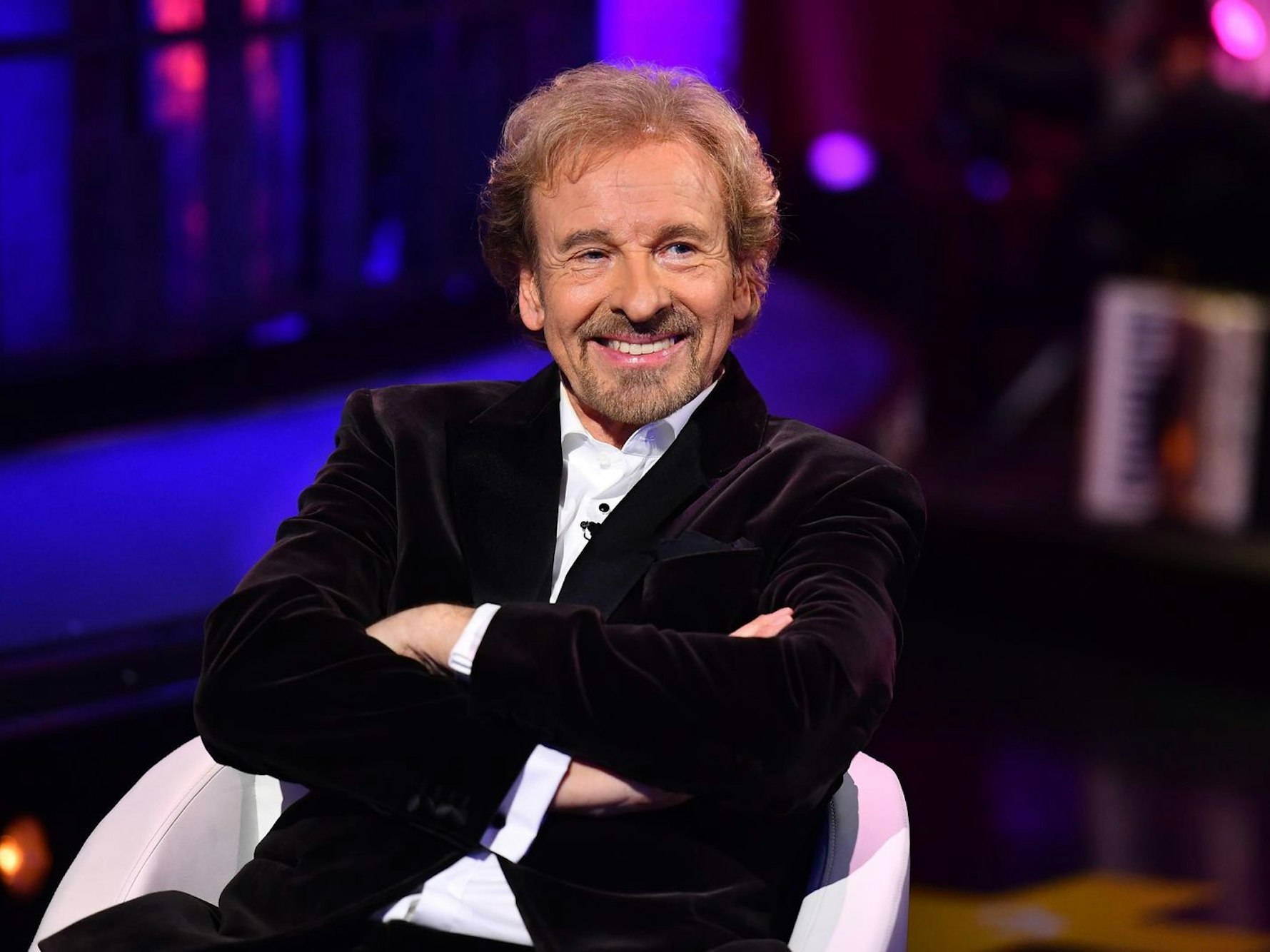 Thomas Gottschalk sitzt mit verschränkten Armen im TV-Studio und lächelt.