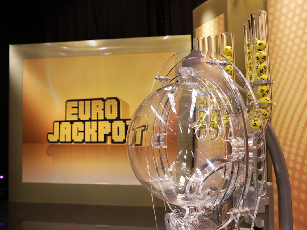 Das Bild zeigt die moderne Ziehungsmaschine für die Eurojackpot-Lotterie mit gelben Kugeln im Vordergrund. Im Hintergrund ist groß das goldene „EURO JACKPOT“-Logo auf einer beleuchteten Wand zu sehen.