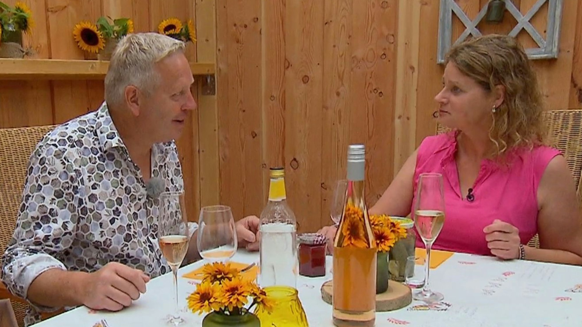 Bauer Jörg und Hofdame Caola sitzen vor gefüllten Sektgläsern an einem Tisch und reden miteinander.