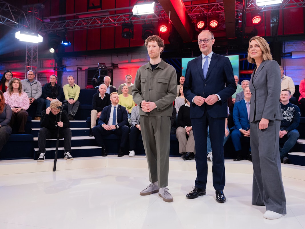 Bundeskanzler Merz steht zwischen zwei Moderatoren inmitten einer Arena, in der das Publikum sitzt.