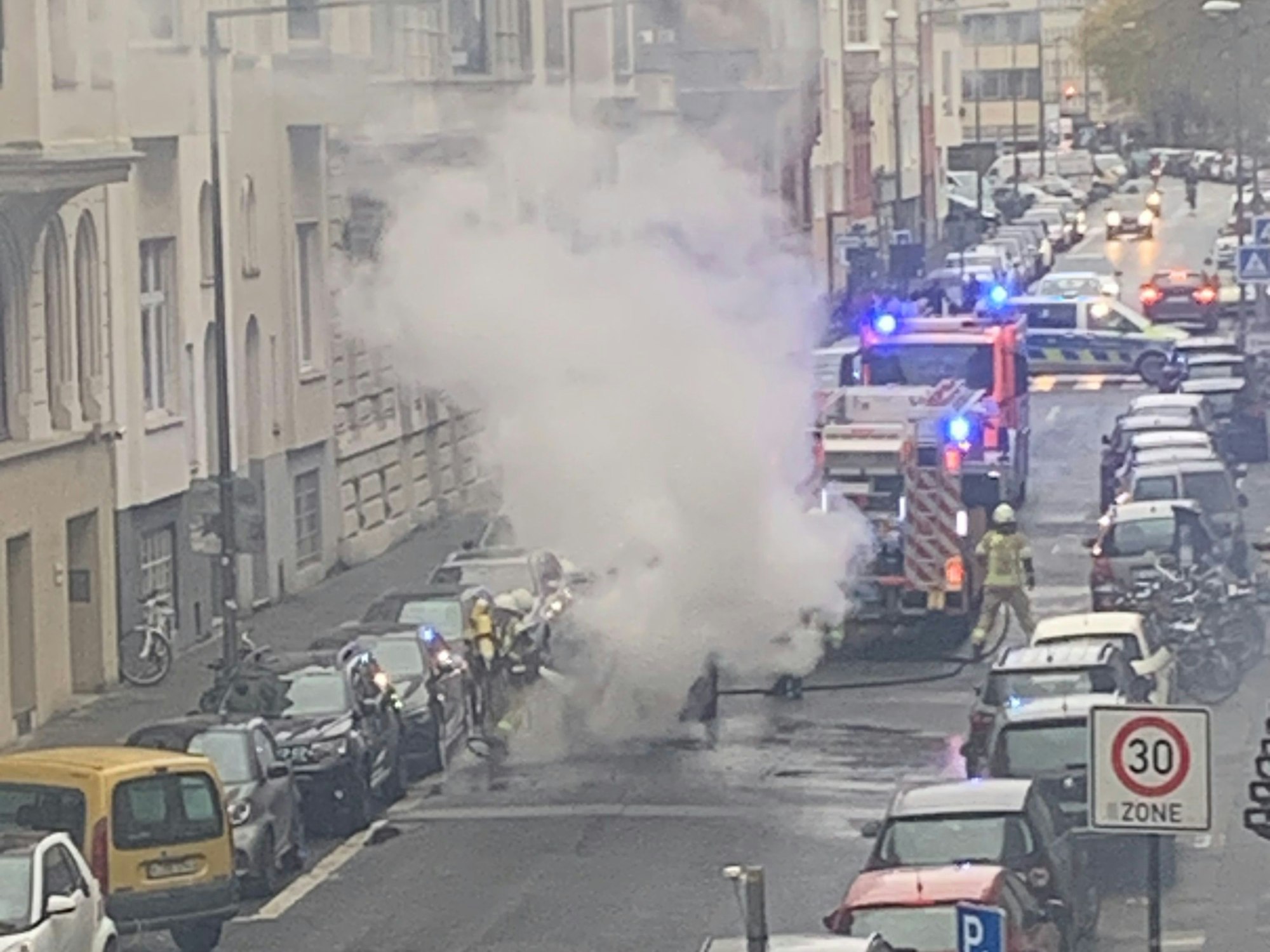 Ein Auto steht auf der Straße und qualmt, Feuerwehrleute löschen.
