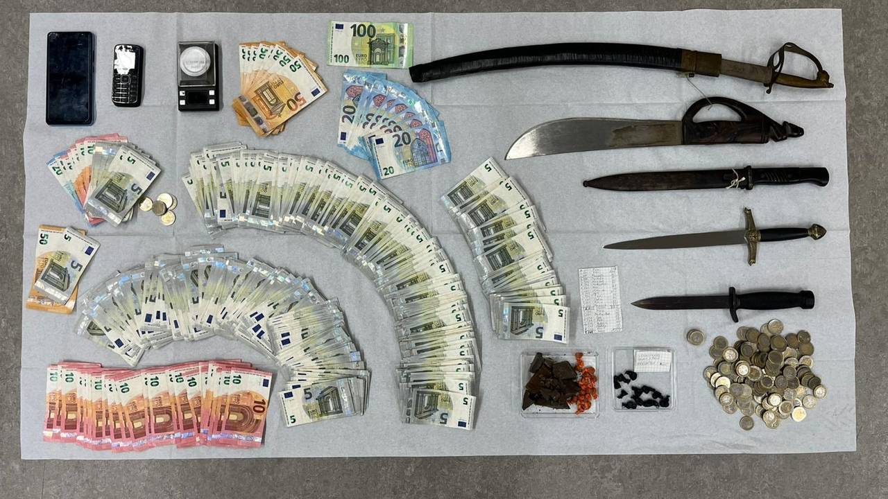 Auf einem Tisch liegen ausgebreitete Geldscheine und mehrere Waffen, darunter zwei Dolche.