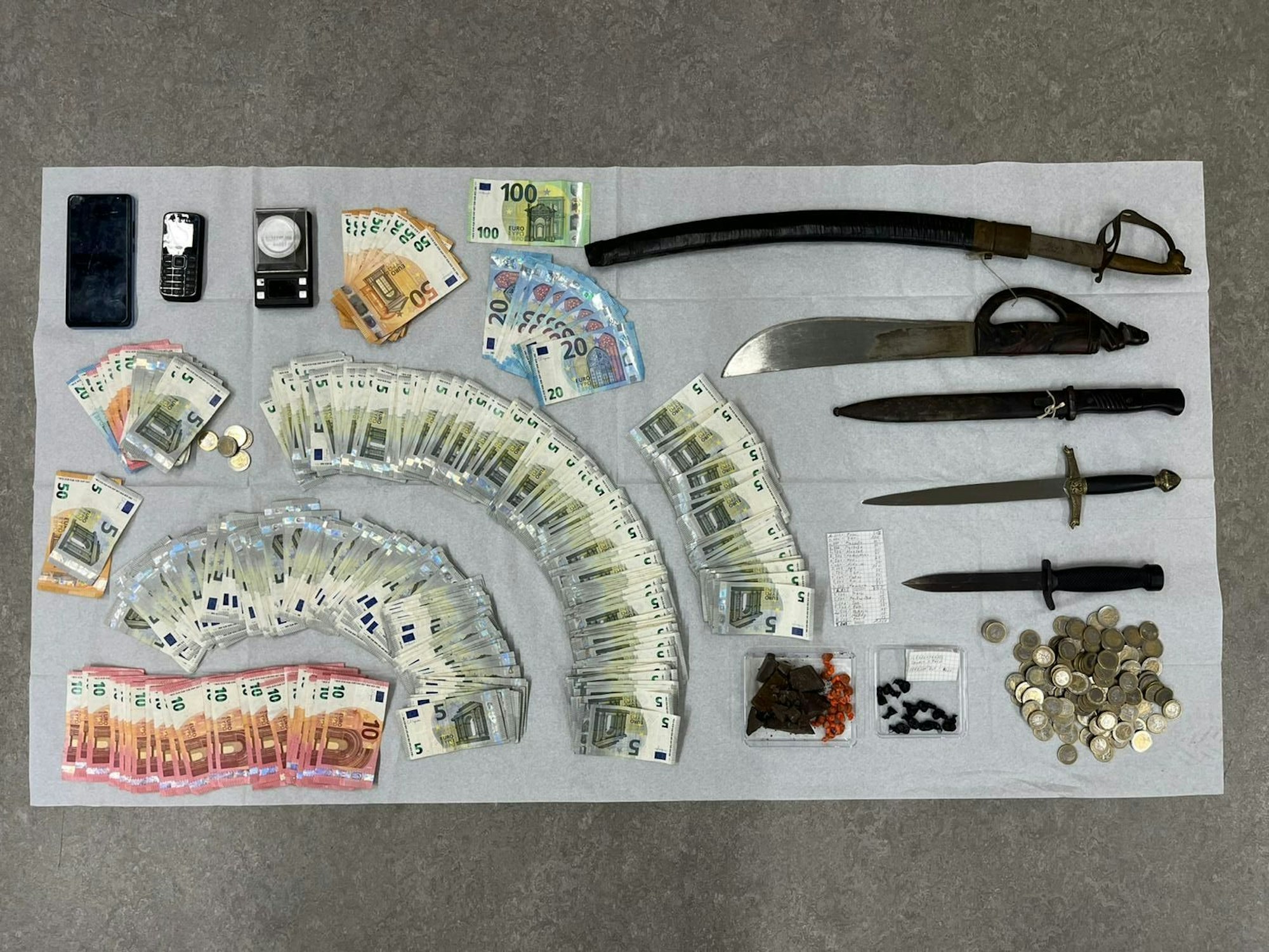 Auf einem Tisch liegen ausgebreitete Geldscheine und mehrere Waffen, darunter zwei Dolche.