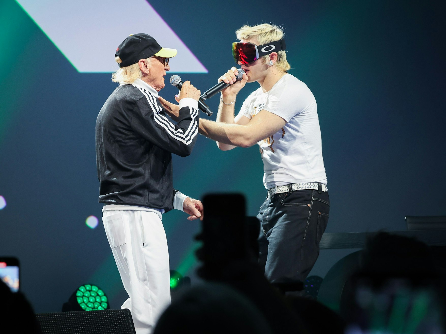 Zwei Sänger, ein älterer Mann mit Sportjacke und Baseballmütze sowie ein jüngerer mit blondierten Haaren und einer Skibrille, stehen sich auf einer Bühne gegenüber und singen sich an.