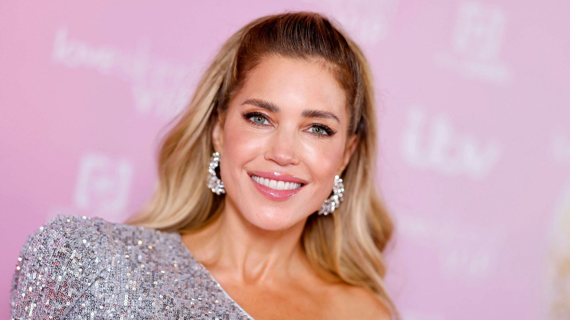 Sylvie Meis lächelt in funkelndem Oberteil vor rosa Hintergrund.
