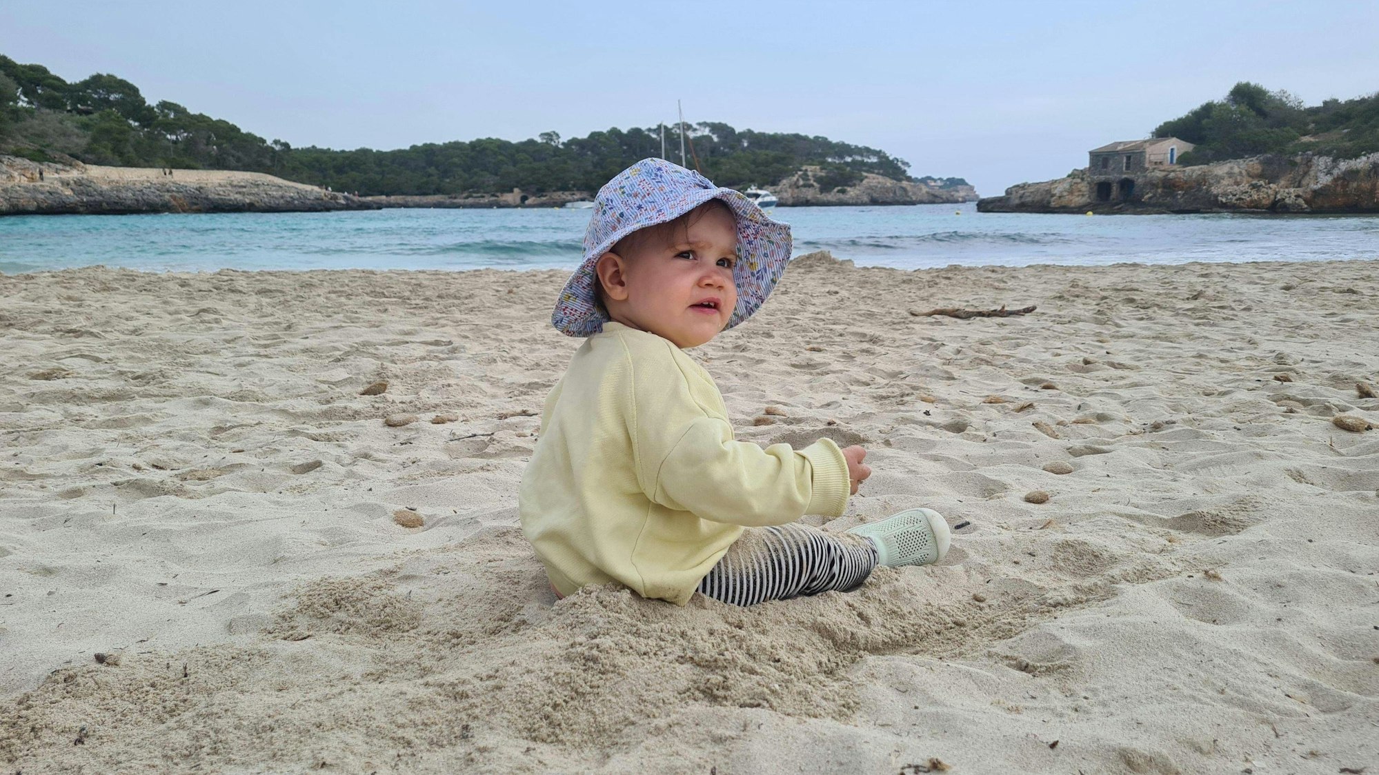 Ein Kleinkind mit Sonnenhut sitzt am Sandstrand am blauen Meer (Symbolbild).