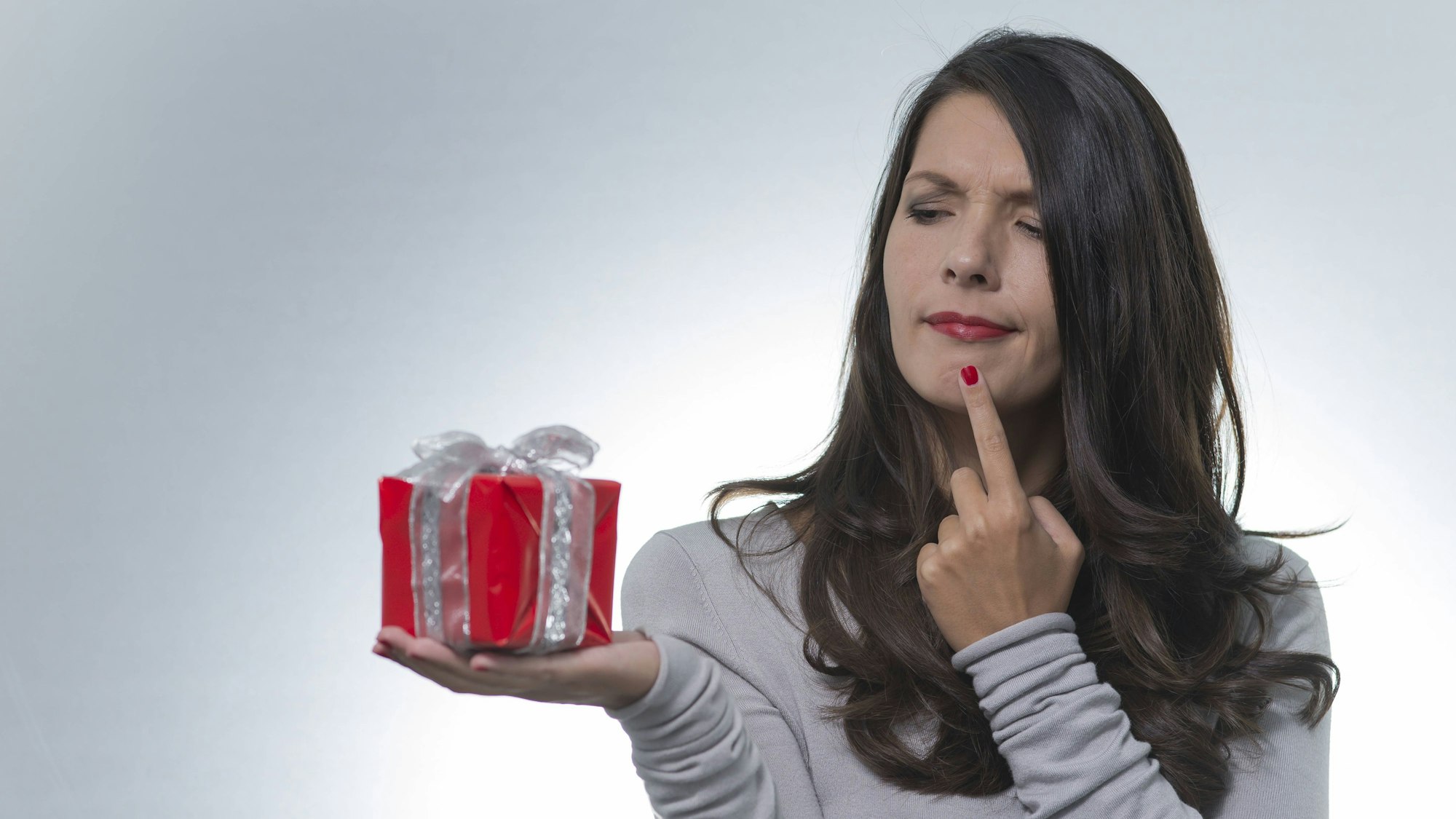 Eine junge Frau mit langen brünetten Haaren, hält ein Geschenk auf der Handfläche und guckt es verwirrt an.