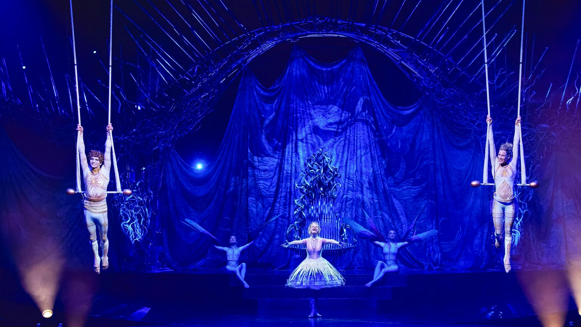 Artisten in weißen Kostümen auf einer blau ausgeleuchteten Bühne.
