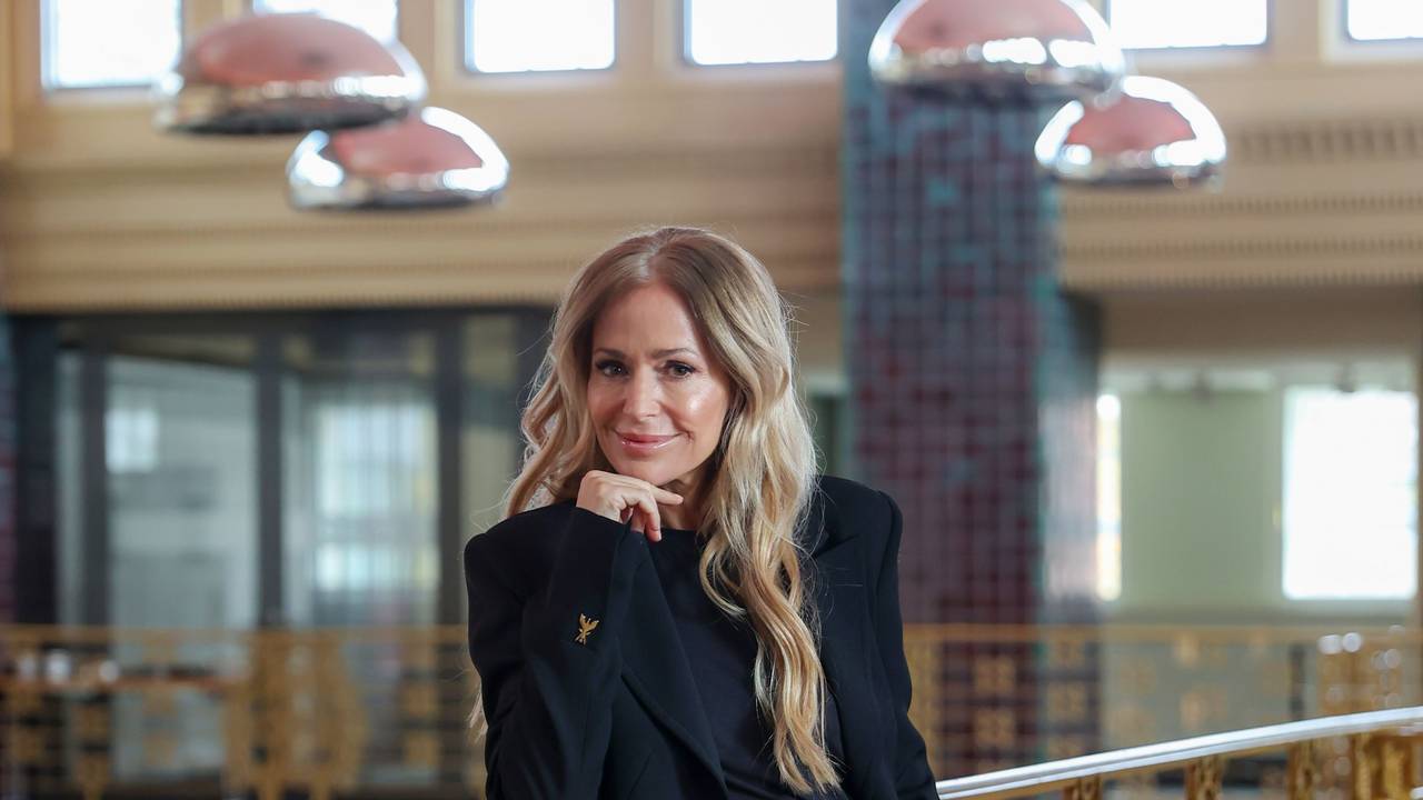 Eine sorgsam geschminkte Frau mit langen blonden Haaren lehnt lächelnd an einem Balkongitter und hat den Kopf auf ihre rechte Hand gestützt.