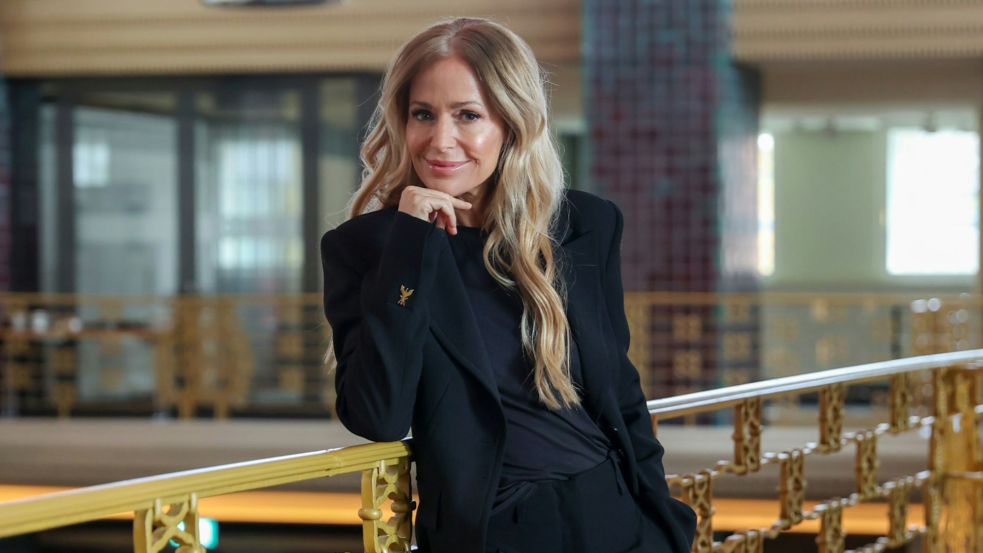 Eine sorgsam geschminkte Frau mit langen blonden Haaren lehnt lächelnd an einem Balkongitter und hat den Kopf auf ihre rechte Hand gestützt.