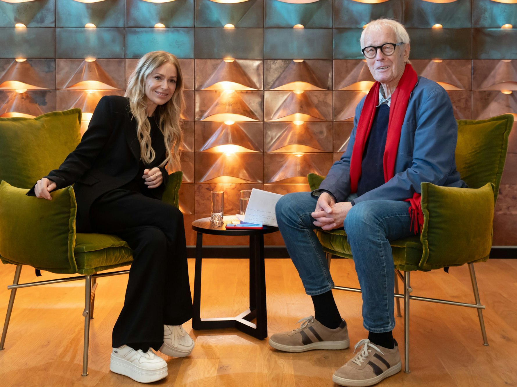 Eine junge Frau mit langen blonden Haare in einem schwarzen Hosenanzug sitzt einem älteren Herrn mit blauem Sakko und rotem Schal gegenüber. Beide sitzen vor einer golden angestrahlten Wand und lächeln.
