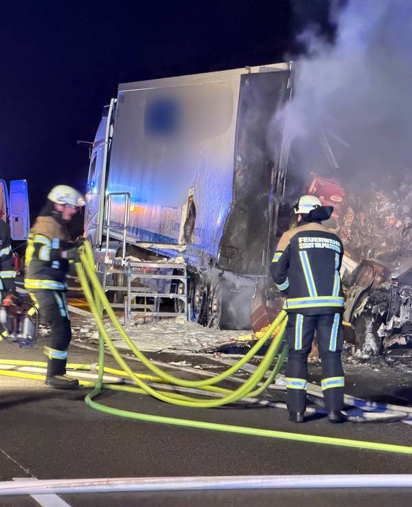 Ein ausgebrannter Pkw steht hinter einem halb ausgebrannten Lkw, davor sind zwei Feuerwehrleute im Einsatz.