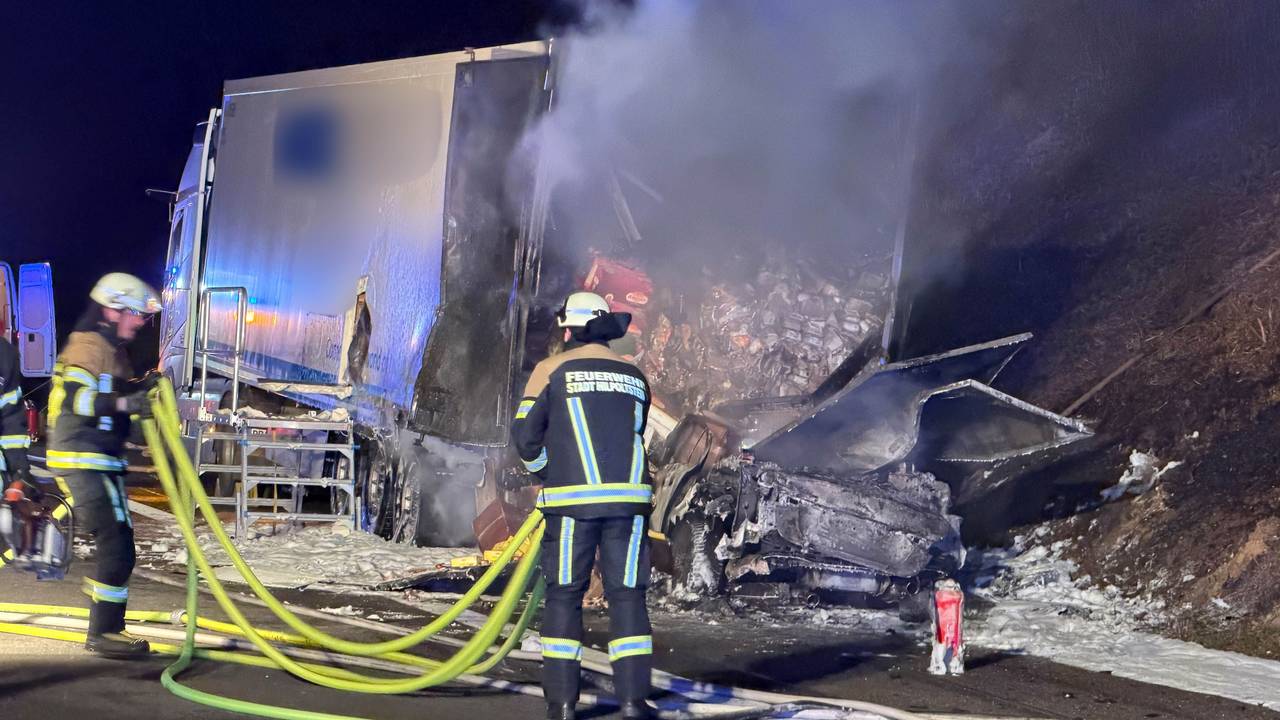 Ein ausgebrannter Pkw steht hinter einem halb ausgebrannten Lkw, davor sind zwei Feuerwehrleute im Einsatz.
