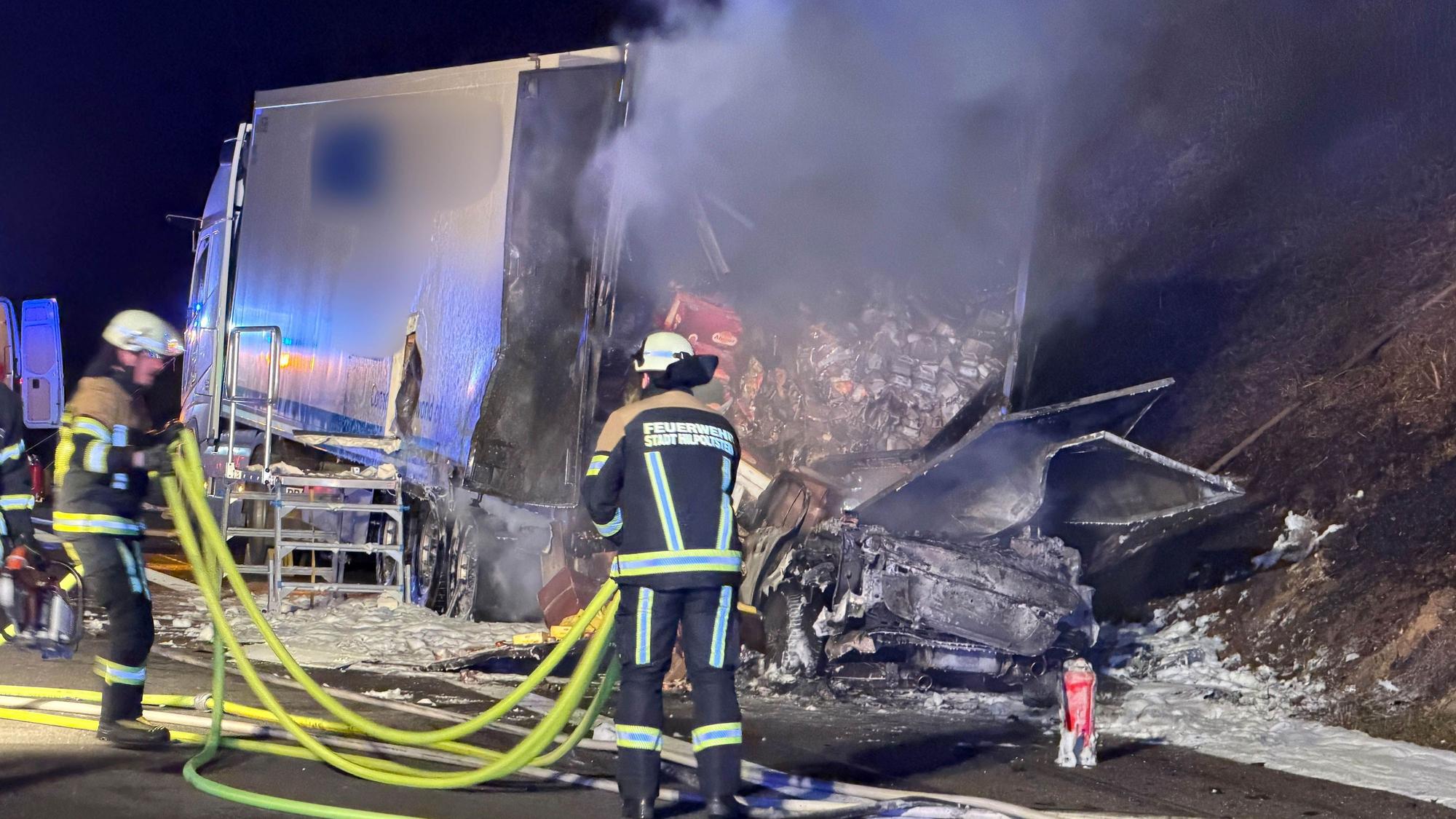 Ein ausgebrannter Pkw steht hinter einem halb ausgebrannten Lkw, davor sind zwei Feuerwehrleute im Einsatz.