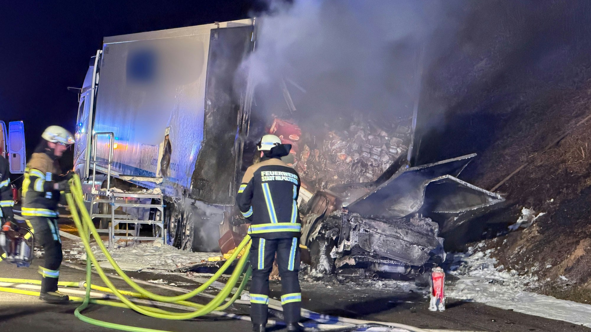 Ein ausgebrannter Pkw steht hinter einem halb ausgebrannten Lkw, davor sind zwei Feuerwehrleute im Einsatz.