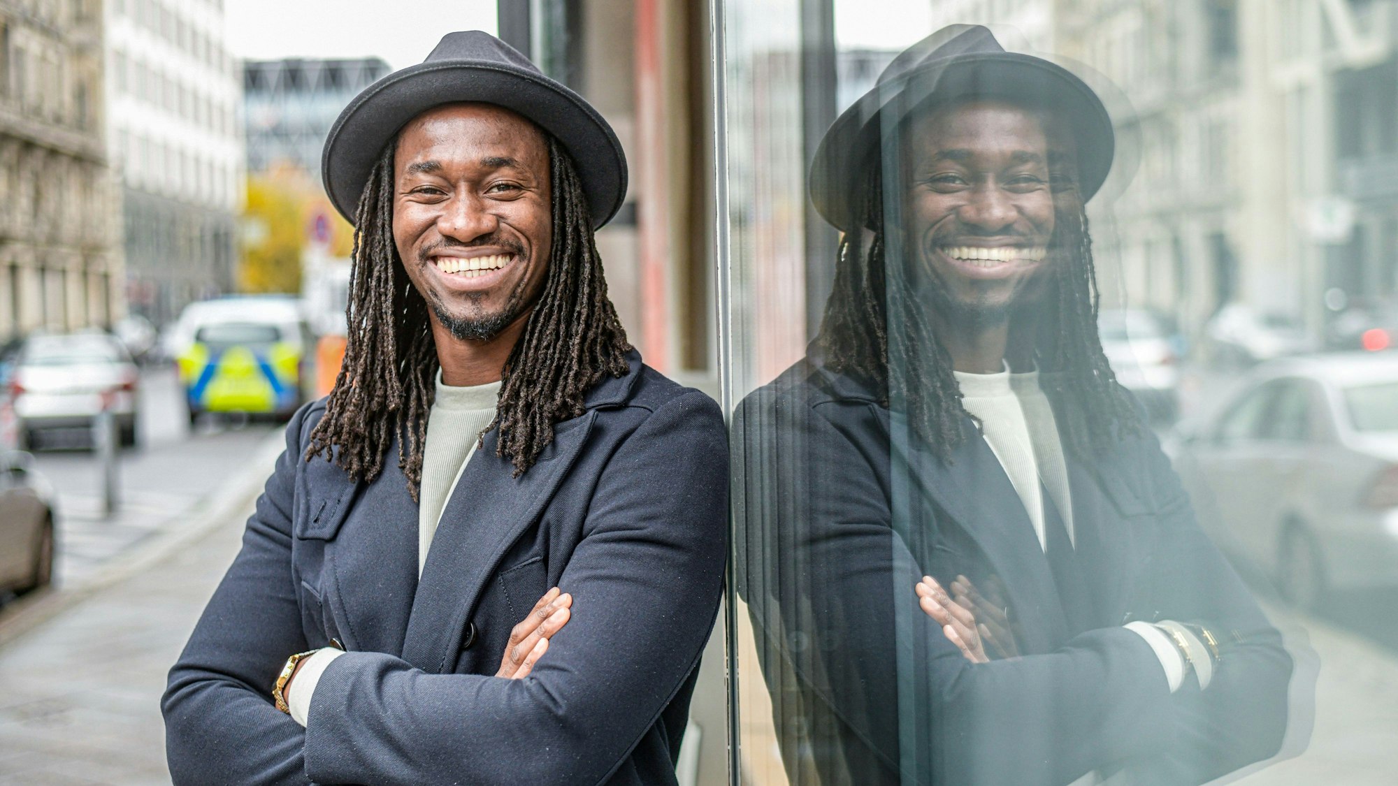 Ein Mann mit Hut und schwarzen Rastazöpfen steht auf einer Straße und lacht fröhlich in die Kamera