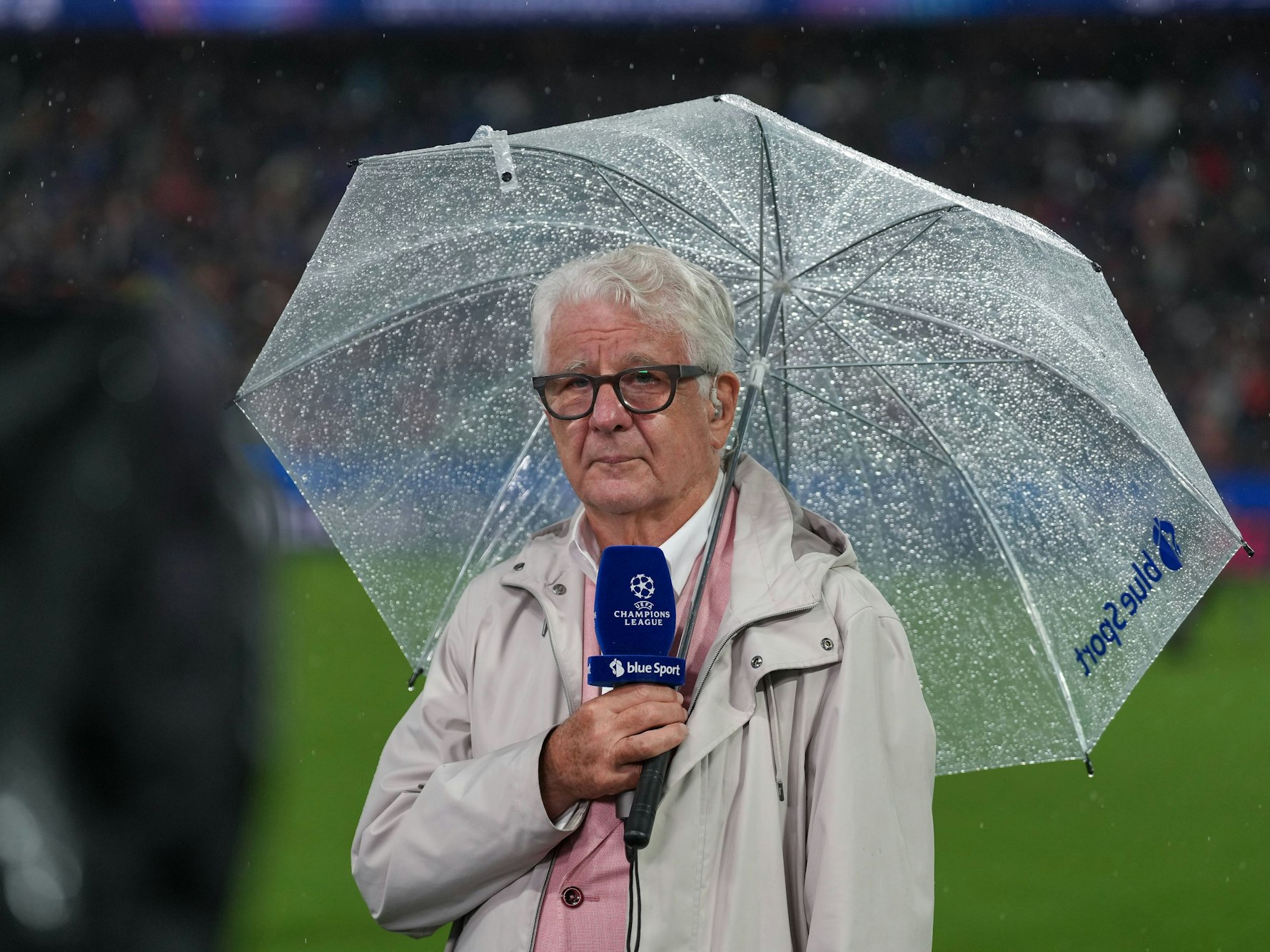 Marcel Reif hat einen durchsichtigen Regenschirm über sich und hält ein blaues Champions-League-Mikrofon in der Hand.