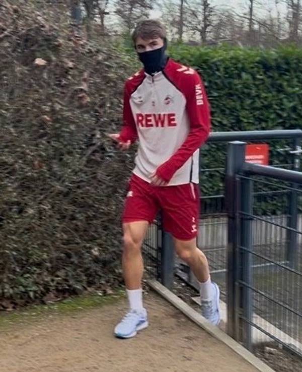 Joel Schmied geht in Trainingsklamotten am Geißbockheim aus einem Tor heraus und hat einen Schlauchschal an, der sein Gesicht bedeckt.