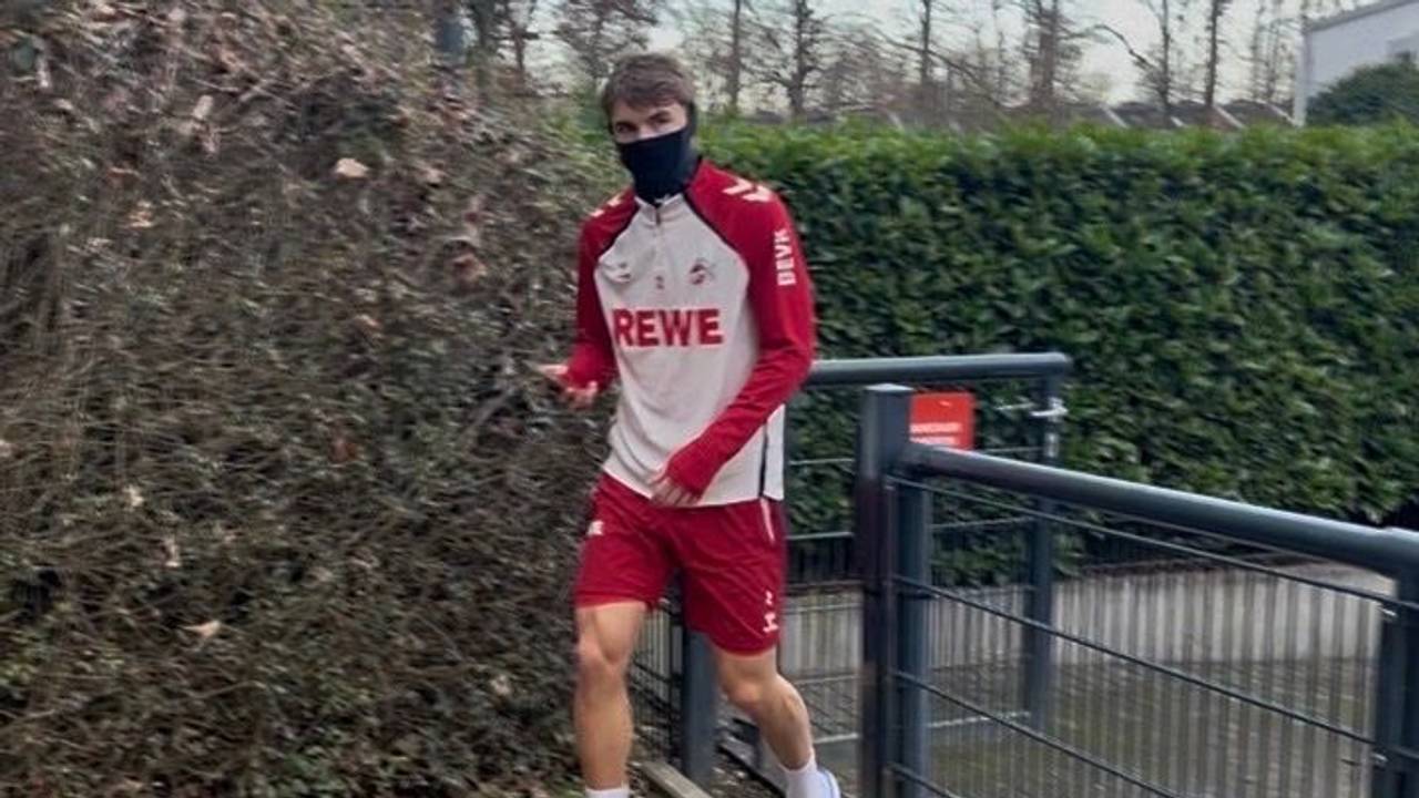 Joel Schmied geht in Trainingsklamotten am Geißbockheim aus einem Tor heraus und hat einen Schlauchschal an, der sein Gesicht bedeckt.