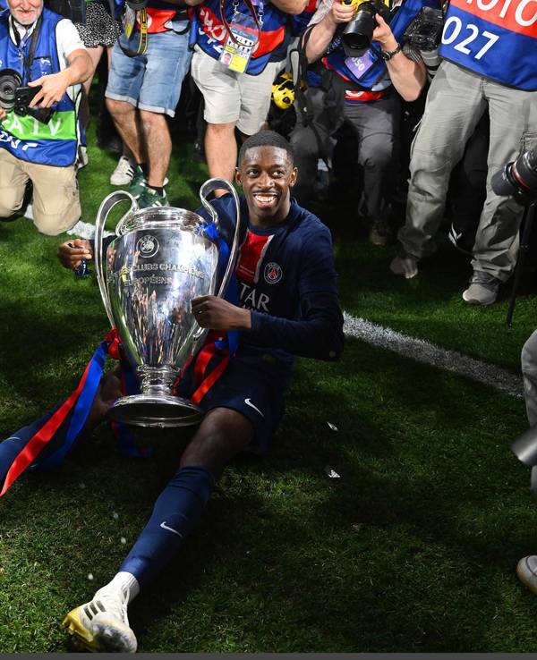 Dembélé sitzt mit der Champions-League-Trophäe auf dem Boden und lächelt.