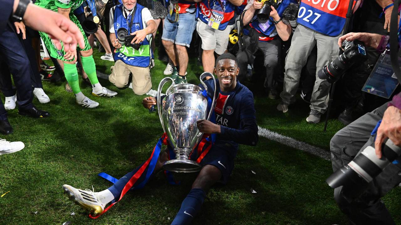 Dembélé sitzt mit der Champions-League-Trophäe auf dem Boden und lächelt.