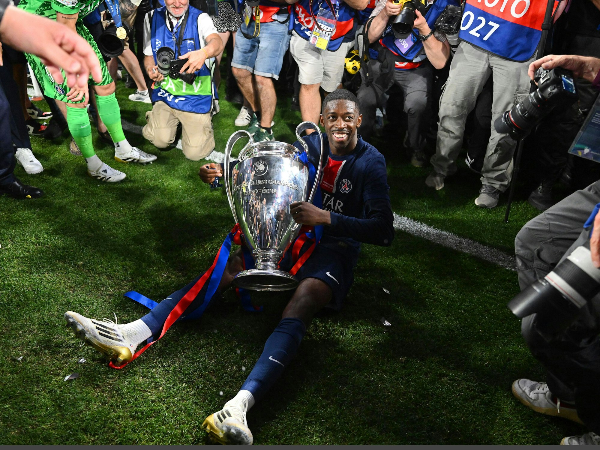 Dembélé sitzt mit der Champions-League-Trophäe auf dem Boden und lächelt.