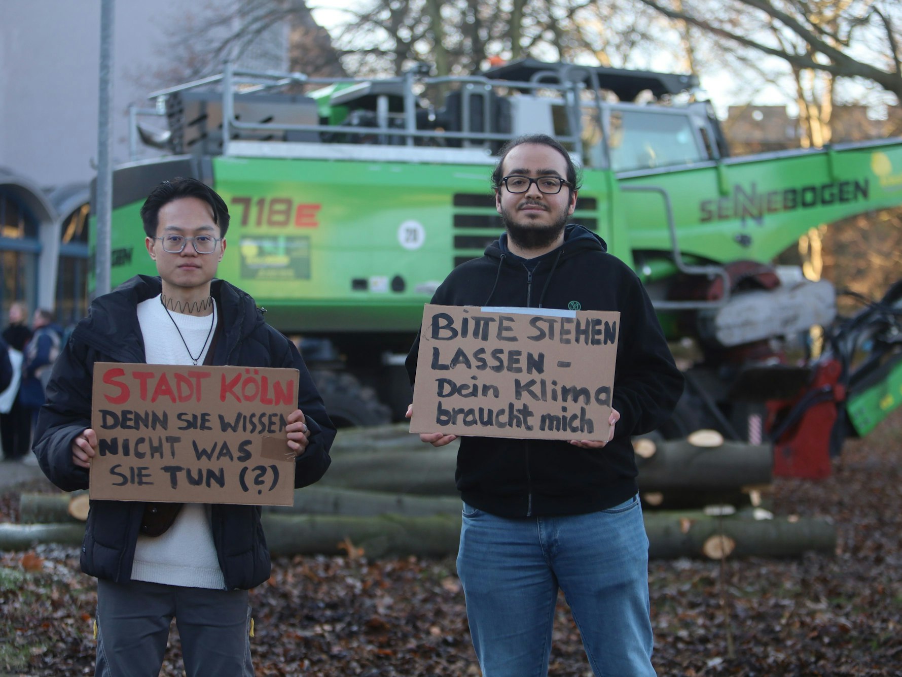 Zwei junge Männer stehen mit Schildern vor einem Bagger, darauf steht "Bitte stehen lassen, dein Klima braucht mich".