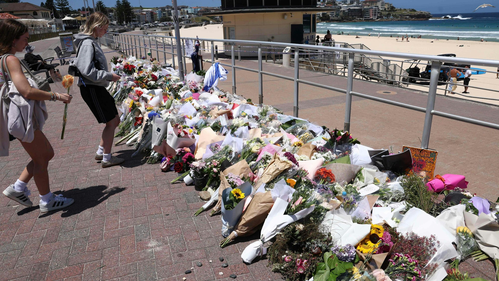 An der Strandpromenade liegen unzählige Blumensträuße in mehreren Reihen, auch zwei Frauen legen Blumen nieder.