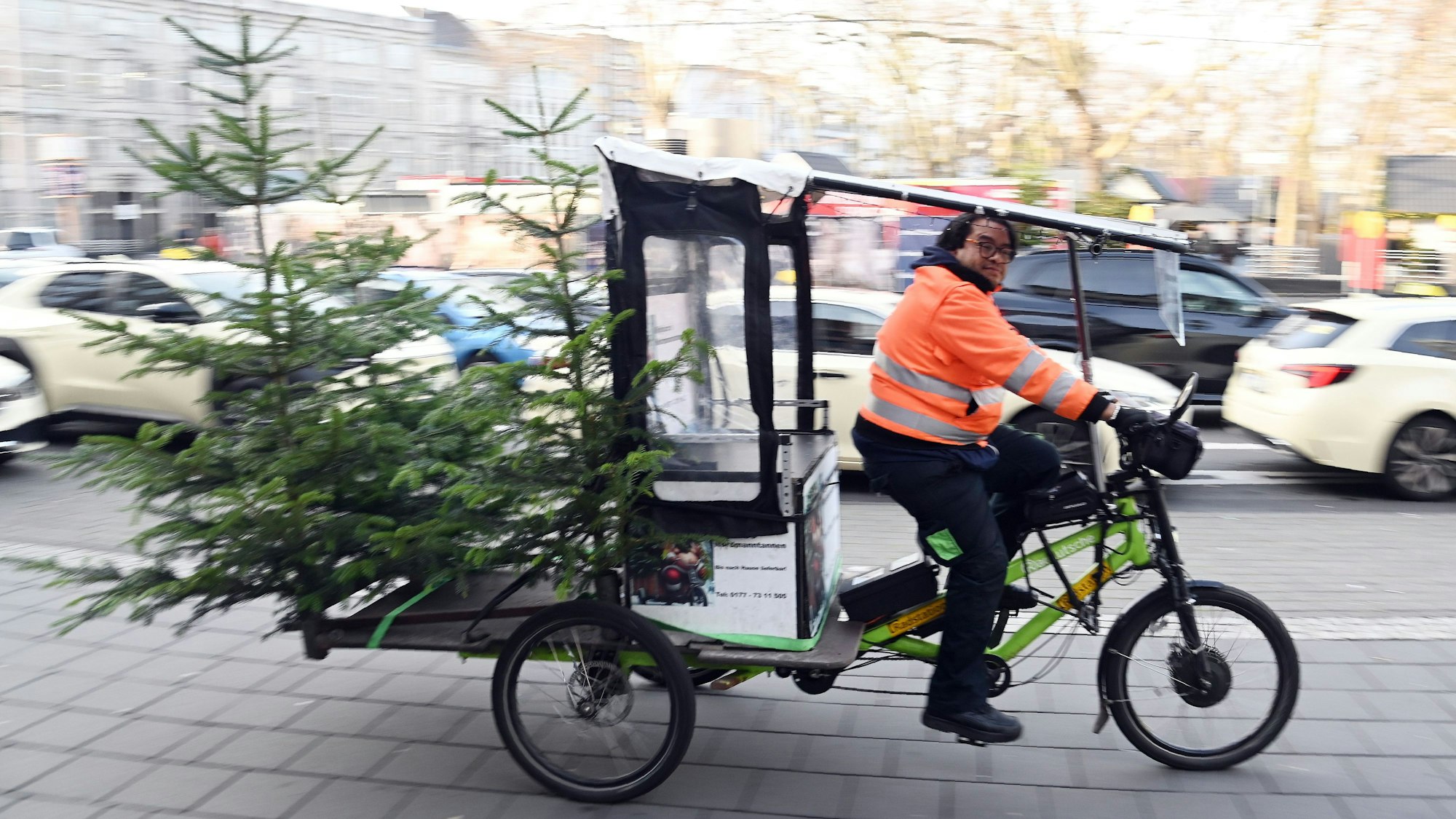 Ein Mann fährt auf einem dreirädrigen Fahrrad mit Ladefläche, auf der zwei Tannenbäume stehen.