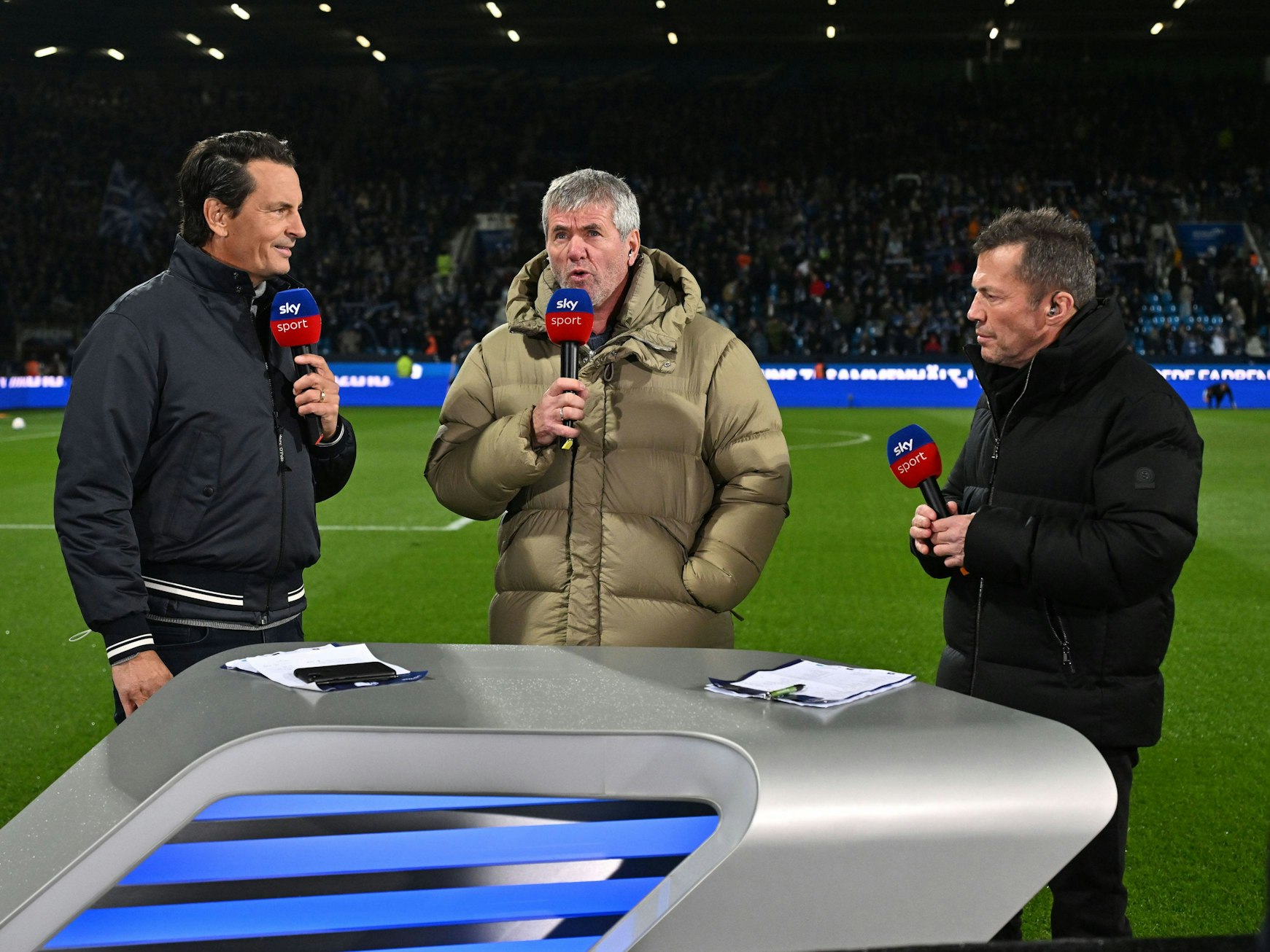 Sebastian Hellmann, Friedhelm Funkel und Lothar Matthäus stehen hinter einem Tisch von Sky und sprechen in Mikrofone.