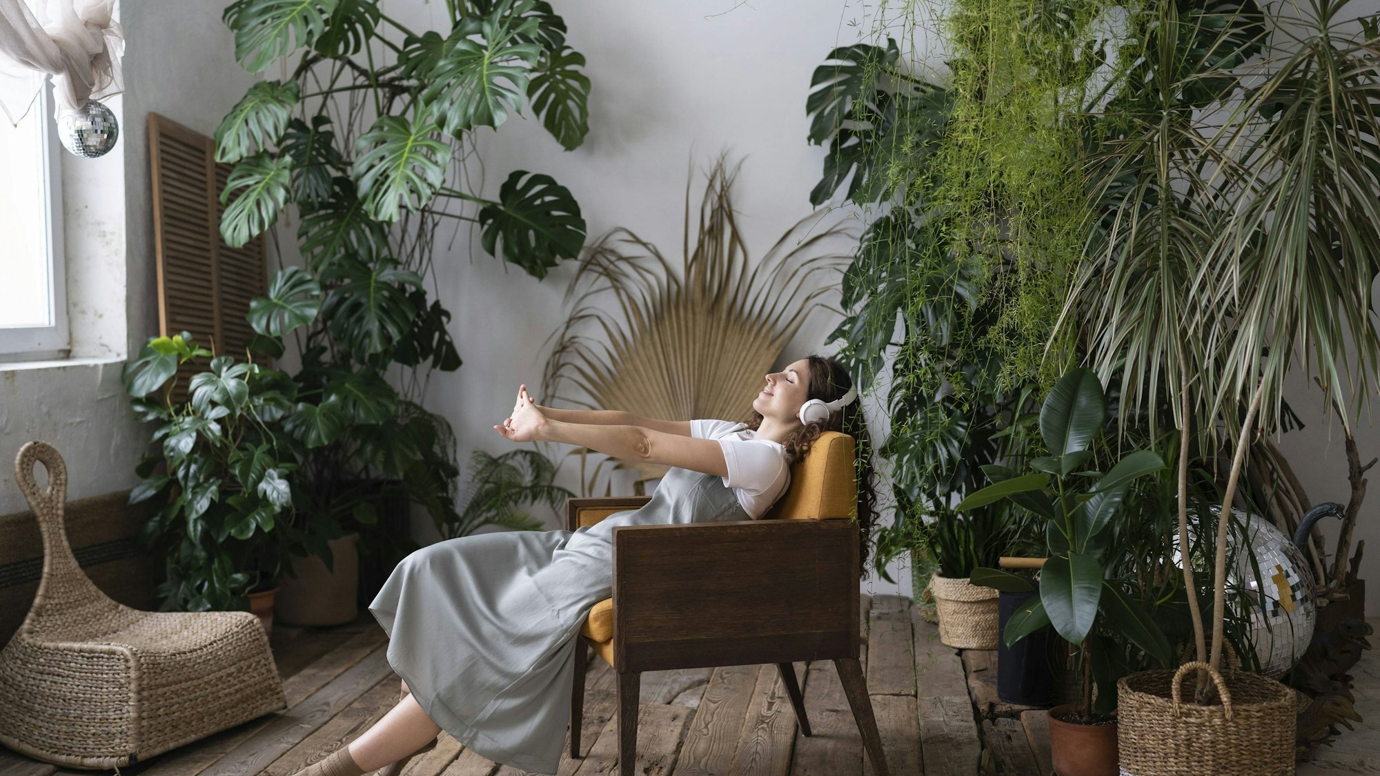 Entspannte junge Frau sitzt inmitten von Grünpflanzen in einem Sessel und streckt sich genüsslich. Sie trägt einen Kopfhörer.