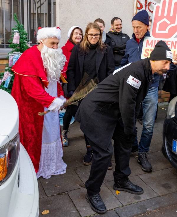 Eine als Weihnachtsmann verkleidete Person schlägt mit einer Rute auf einen Mann ein, an dessen Kleidung und Mütze GAG Immobilien steht.