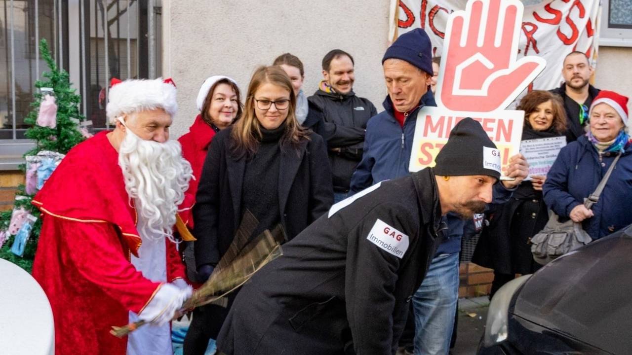 Eine als Weihnachtsmann verkleidete Person schlägt mit einer Rute auf einen Mann ein, an dessen Kleidung und Mütze GAG Immobilien steht.