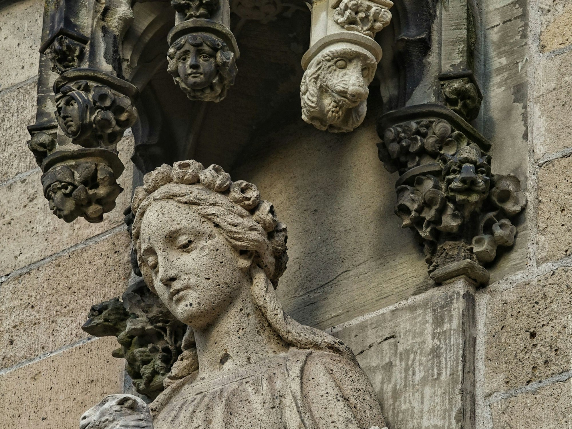 Eine Hundekopf-Skulptur an einem Stein-Baldachin über der Statue einer Frau. An dem Baldachin hänge auch andere Gesichter.