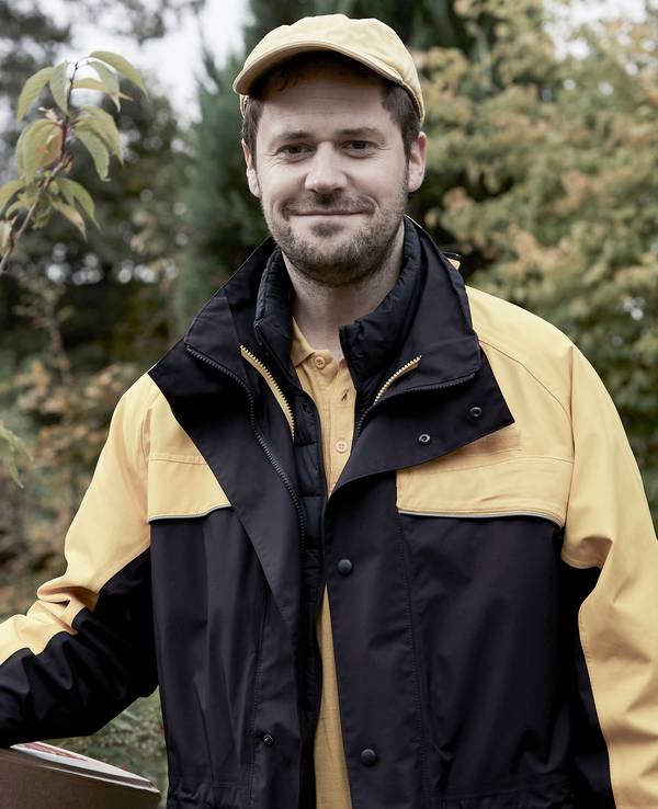 Ein Mann in der Uniform eines Postboten trägt eine gelbe Mütze und lacht in die Kamera