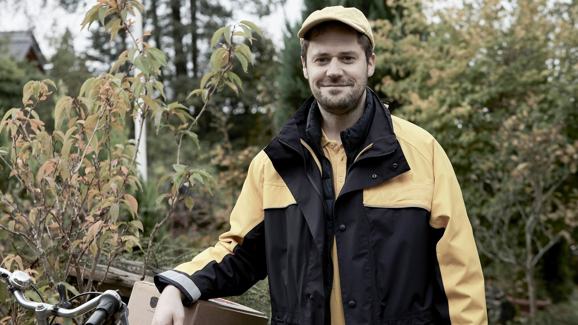 Ein Mann in der Uniform eines Postboten trägt eine gelbe Mütze und lacht in die Kamera