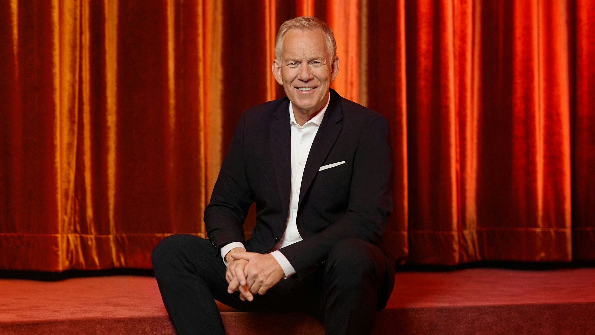 Ein Mann im Anzug mit weißen Sneakern sitzt auf einer roten Showtreppe und lächelt.