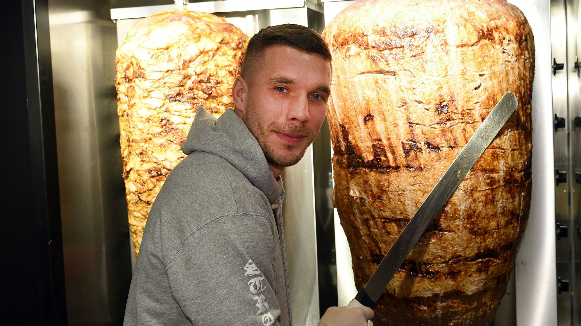 Fußball-Weltmeister Lukas Podolski steht am 6. Januar 2018 in Köln bei der Eröffnung eines Döner-Ladens an einem Dönerspieß.