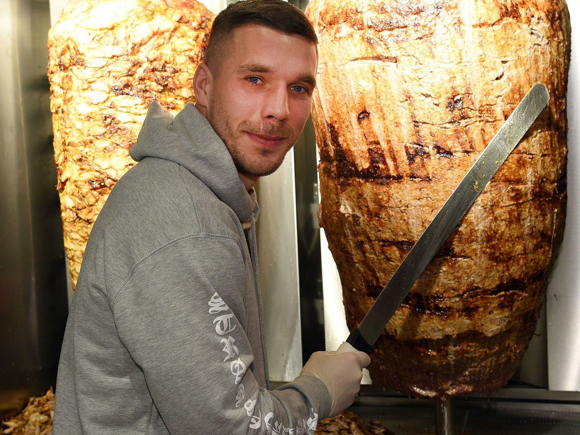 Fußball-Weltmeister Lukas Podolski steht mit einem langen Messer vor einem Dönerspieß.