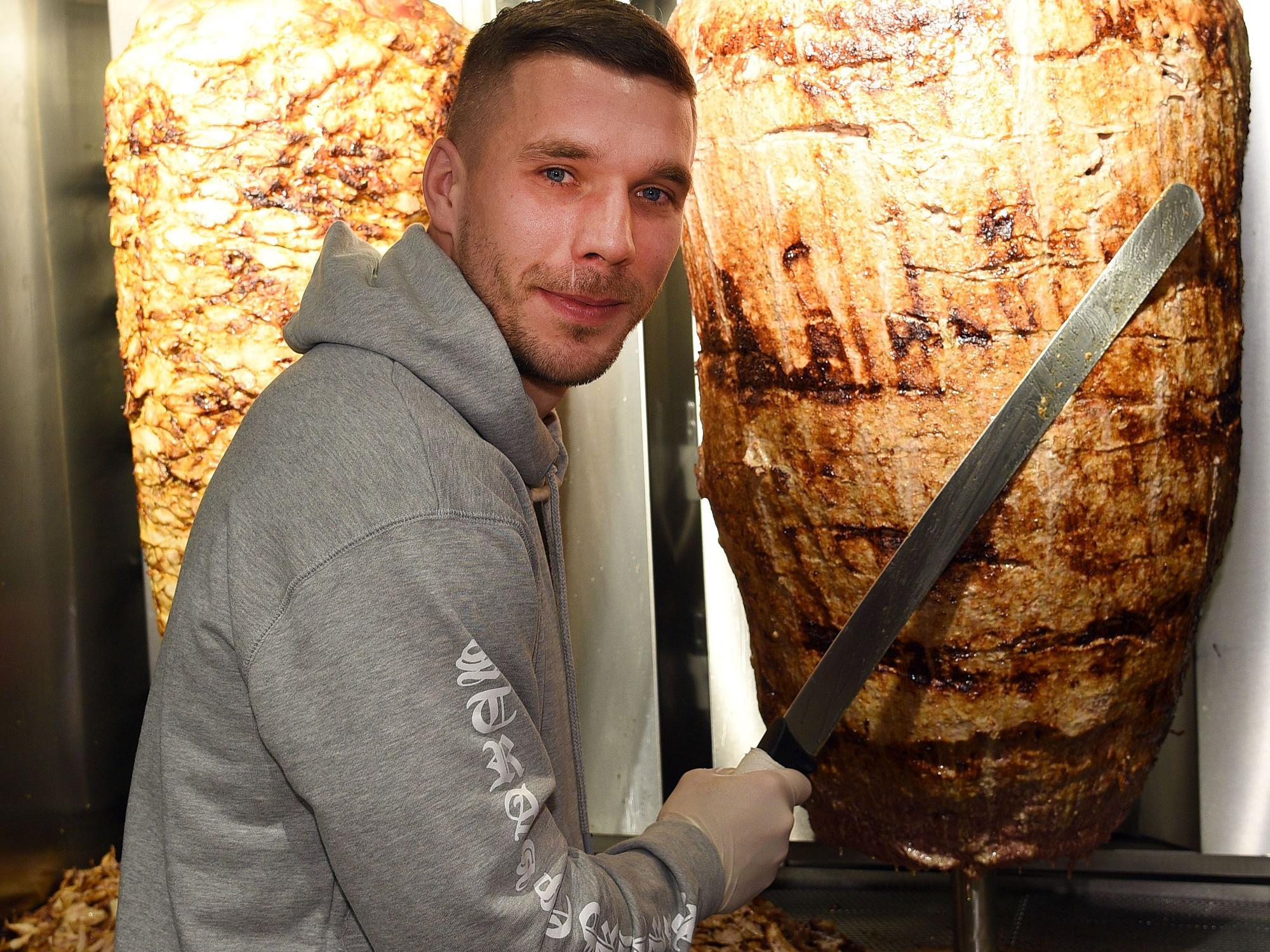 Fußball-Weltmeister Lukas Podolski steht mit einem langen Messer vor einem Dönerspieß.