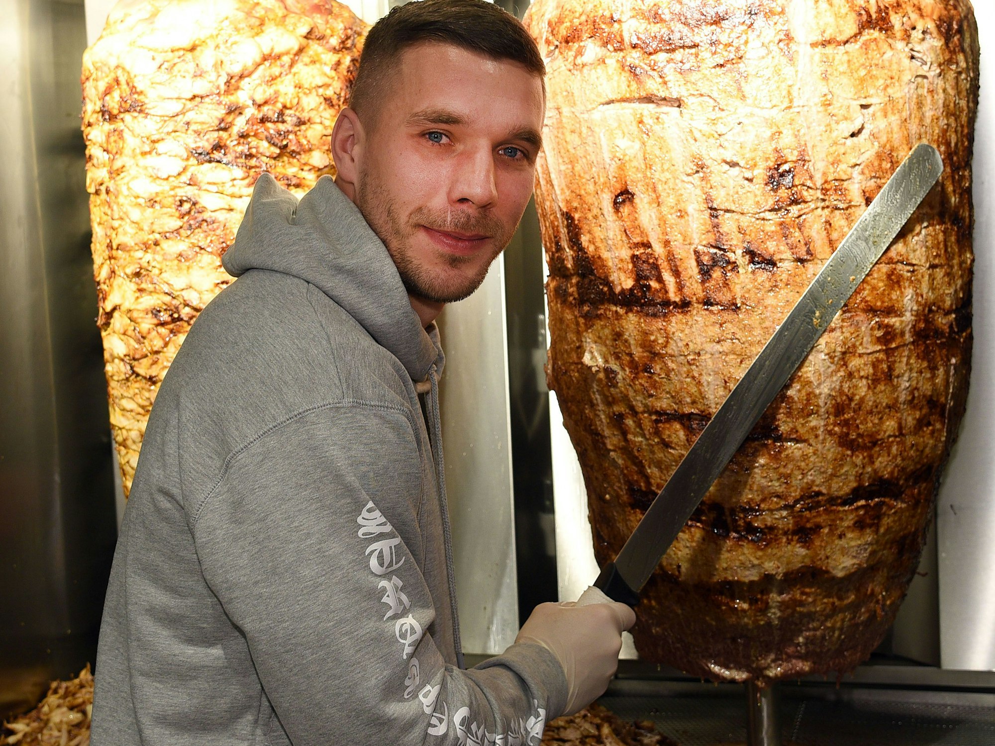 Fußball-Weltmeister Lukas Podolski steht mit einem langen Messer vor einem Dönerspieß.
